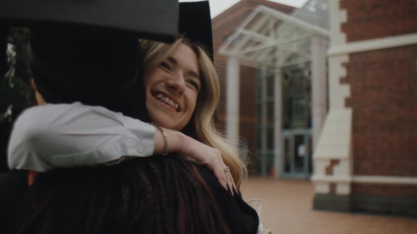 Two college students in graduation gowns greeting each other with a hug and happy
