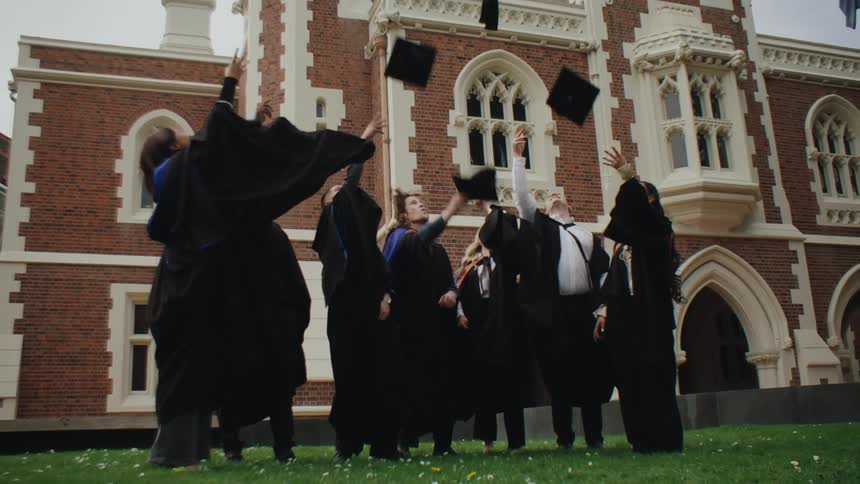 Graduating students stand outside wearing graduation gowns and throwing caps in the air