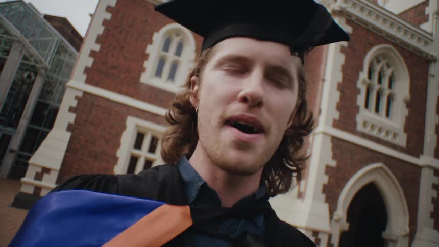 A graduate clad in an academic robe and cap stands at the graduation prom engaging in conversation and scanning the surroundings