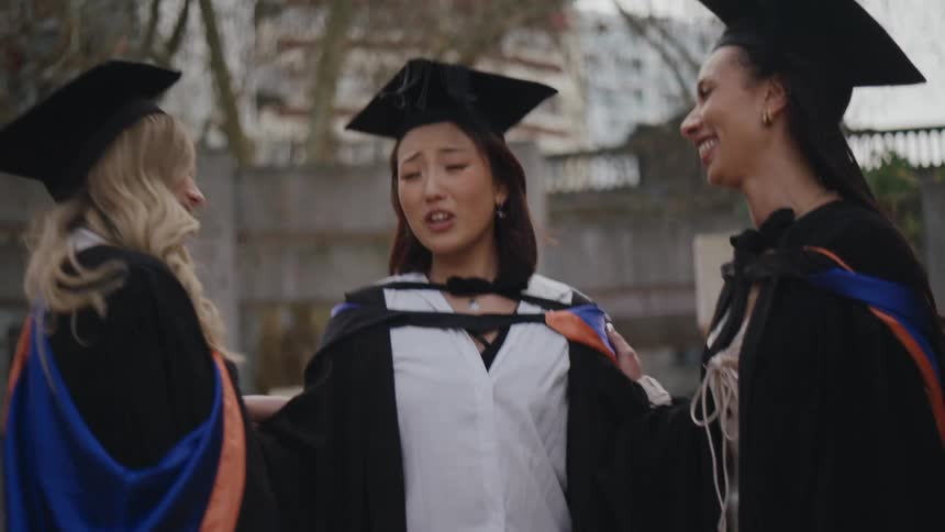 Three women wearing graduation cap and gowns stand outside under tree and chat