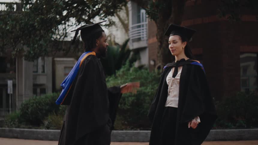 Two individuals in graduation gowns and caps stand outside near a college or university