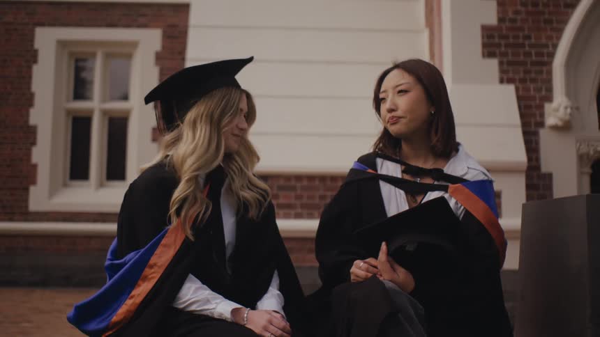 Two graduates in academic robes and caps sit and talk to each other in front of the faculty
