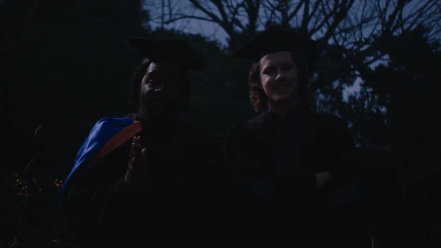 Two graduates in cap and gown one with his arms folded stand outdoors at twilight