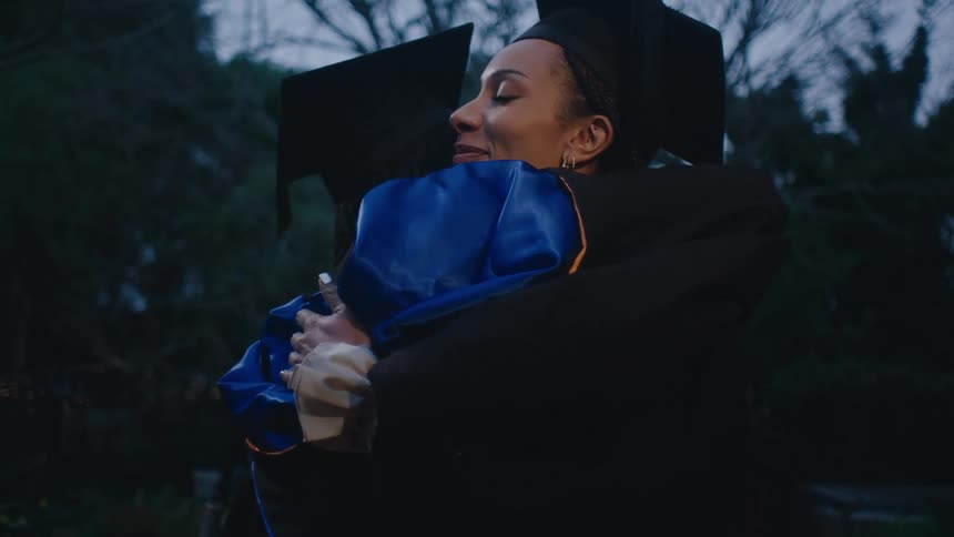 Man and woman wearing graduation clothing hug each other as a light flashes