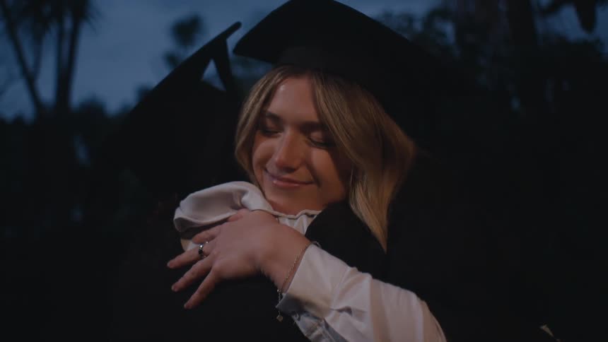 Two graduating students hug and smile outdoors as bright lights illuminate them