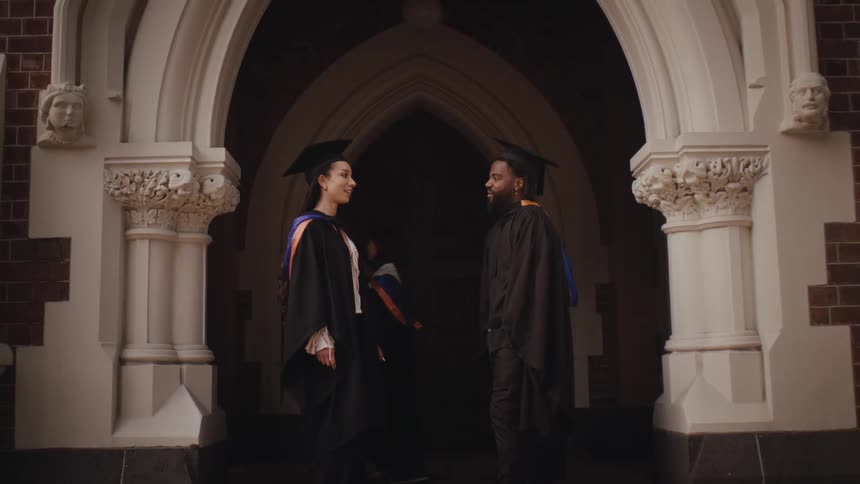 Two people in graduation gowns standing and facing each other under a decorative archway suggesting a university or ceremonial setting