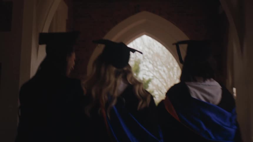 Three individuals wearing graduation caps and gowns walk through an arched doorway with their backs to the camera