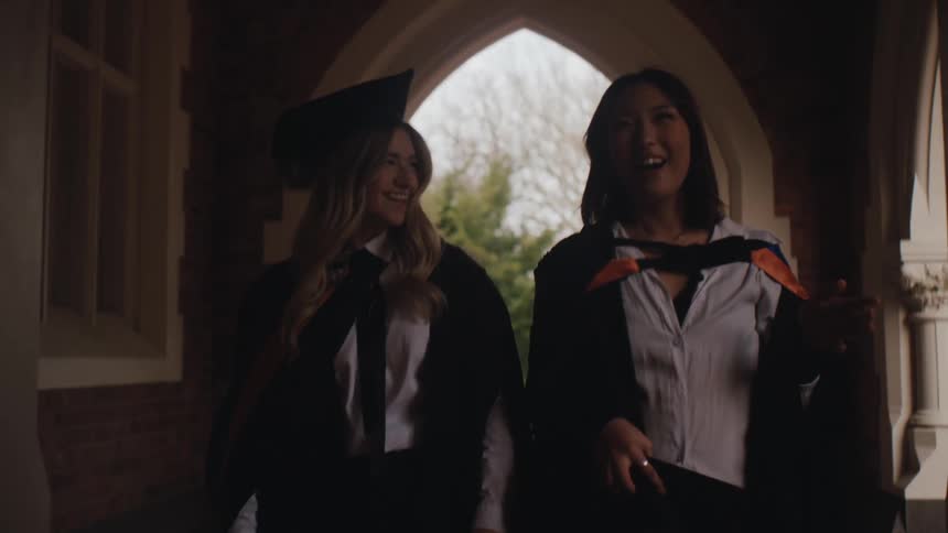 Two graduates in academic robes and caps walk through an arched doorway sharing a smile as they step into the light outside