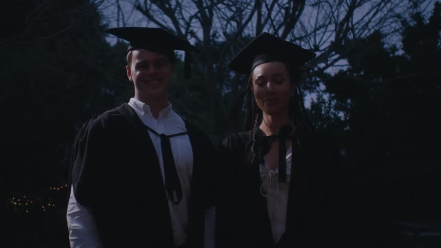 Man and woman standing side by side in graduation gowns and camera flashes appearing
