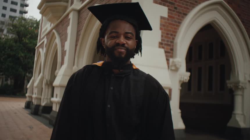A man in a graduation gown and cap stands in front of a building smiling and nodding