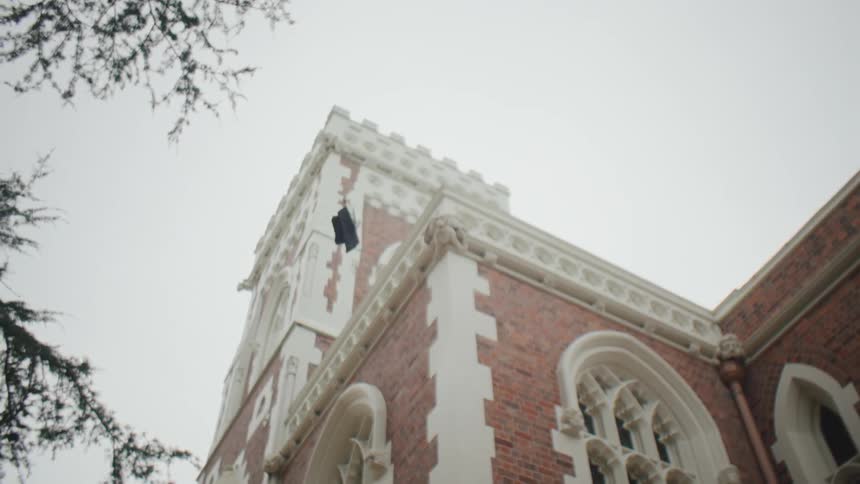 A female student in graduation attire throws her mortarboard into the air outside of a school building