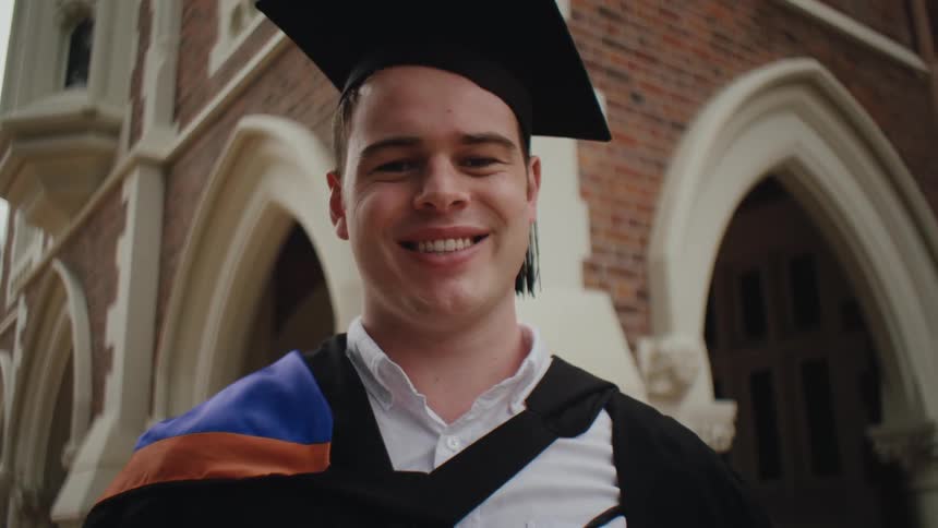 A young man wearing a graduation cap and gown smiles at the camera