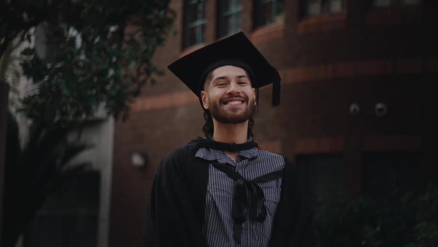 A young man wearing a graduation cap and gown smiles at the camera