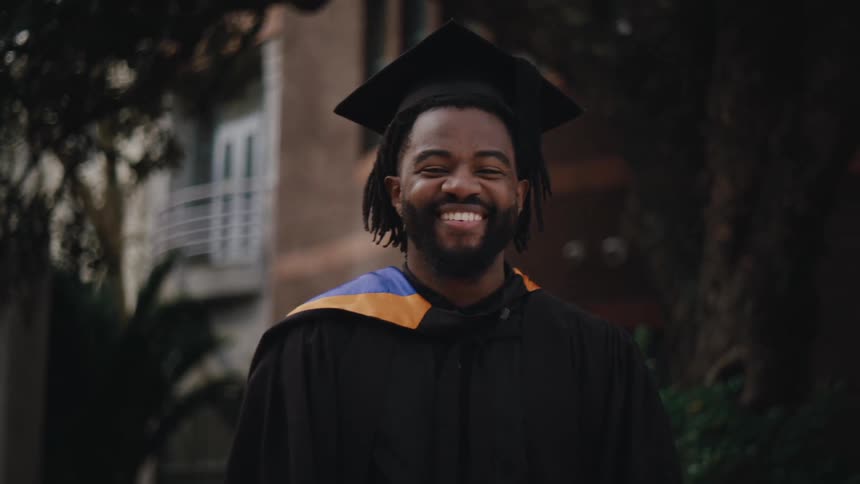 A black man in graduation clothing smiles outside of a school building