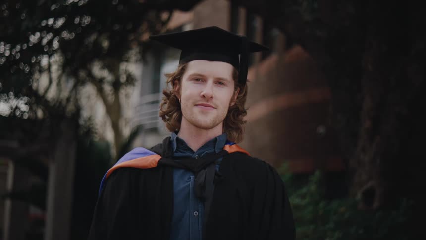 Male graduate smiles while posing near a campus building in his graduate robe and mortarboard