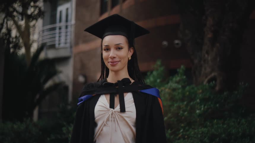 A graduate student in a cap and a gown stands smiling in front of a building