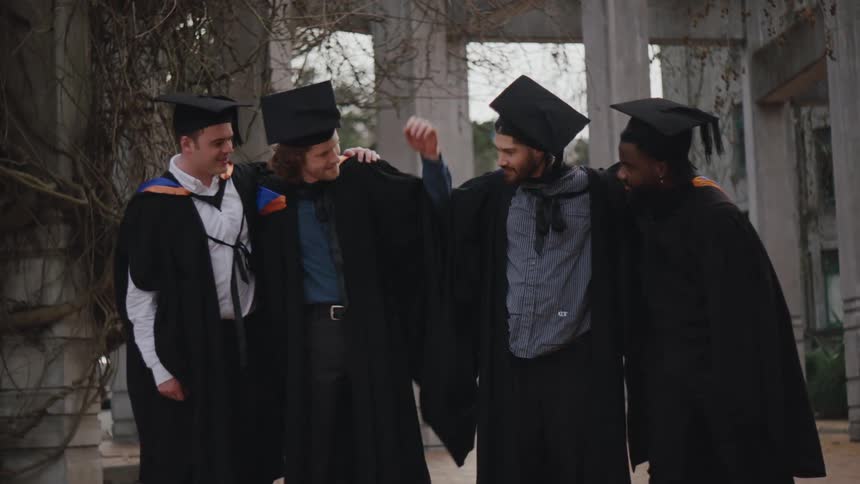 Four people in graduation gowns and caps standing together outdoors celebrating