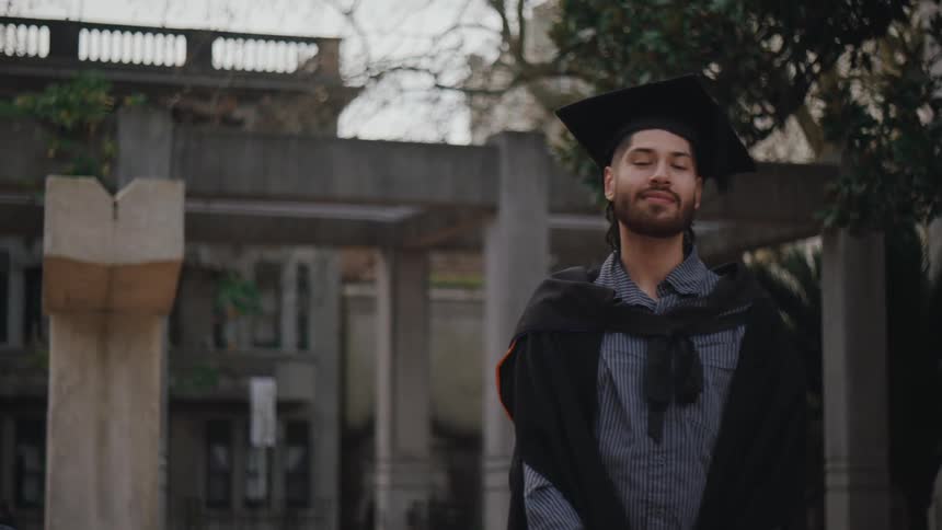 A young man wearing a graduation cap and gown smiles at the camera