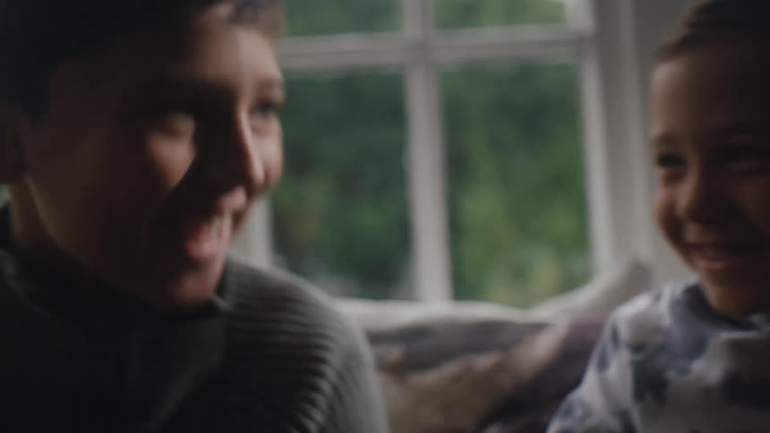Younger brother whispers to his older brother while they sit on the couch in their living room