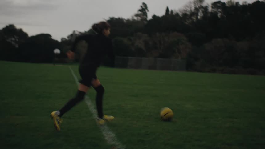 A teen girl on a black uniform with yellow shoes playing soccer on a grassy field