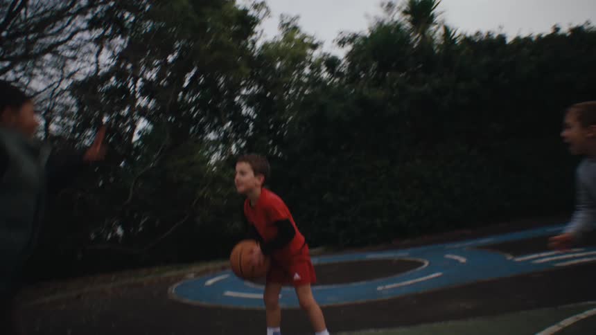 Two children race across an outdoor basketball court with one holding the ball tightly and focused on the game while the others chase closely behind under the dim light of a cloudy sky