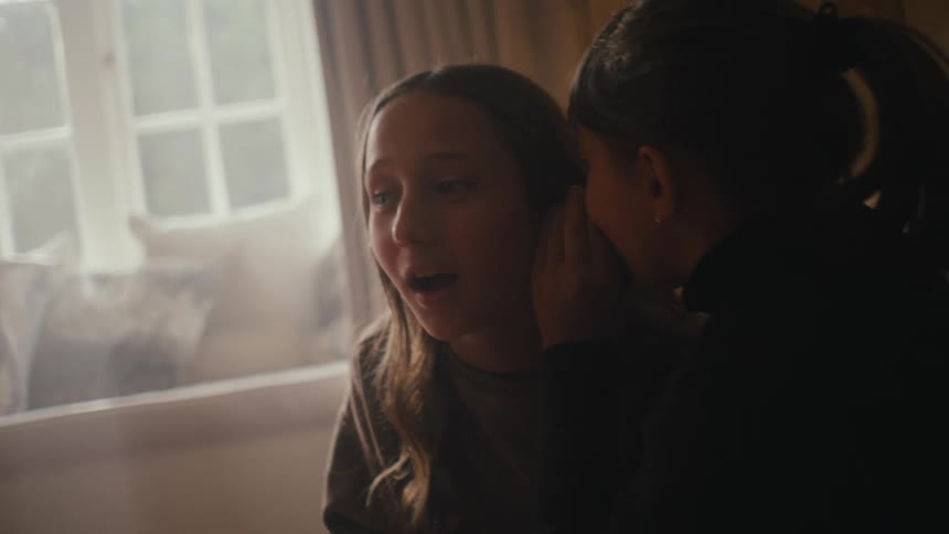 A young girl leans in close to whisper a secret into her friends ear both of them smiling softly in a warmly lit room creating an intimate and playful atmosphere