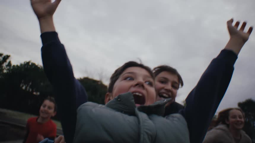 Boy enjoys riding in a cart pushed by his friends through the park past watching adults on an overcast fall day