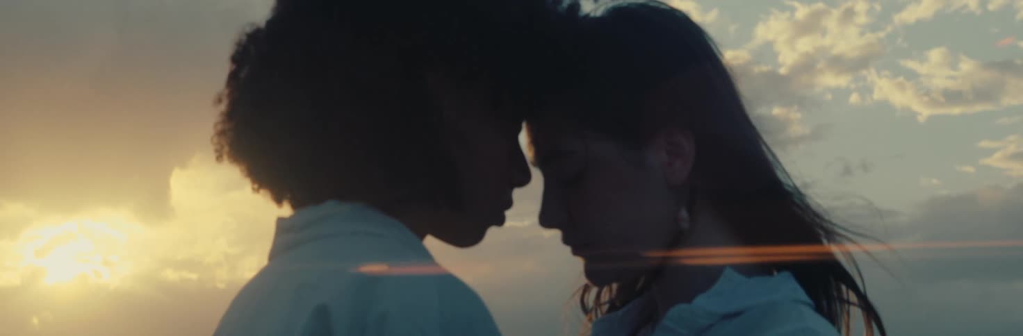 Two people stand close with their foreheads touching against a sunset and cloudy sky background