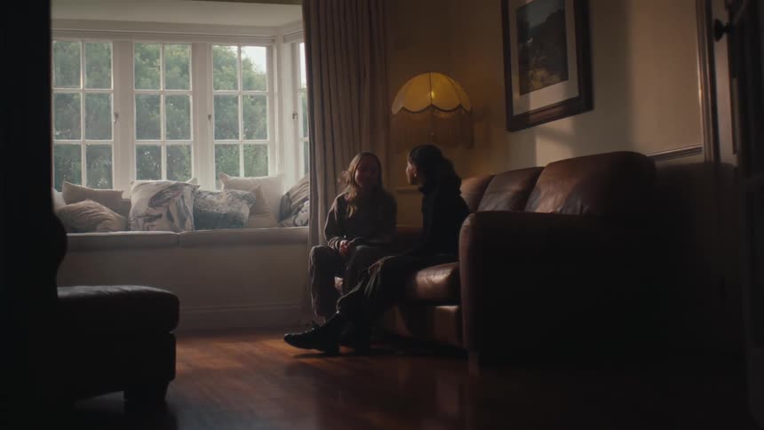 Two people converse in a dimly lit living room with a window seat