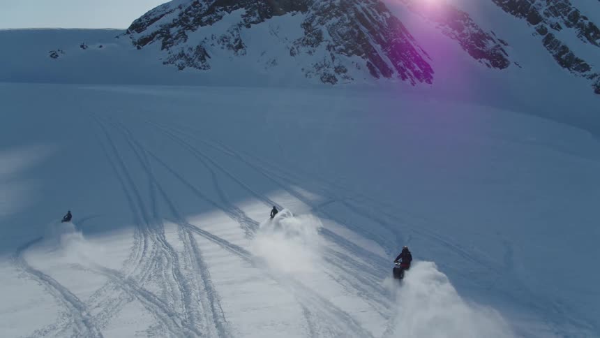 People ride snowmobiles across the snow near the mountains on a sunny winter day