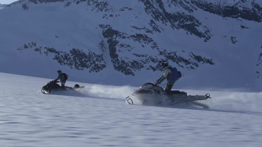 Two people on a snowmobile at high speed with mountains in the background