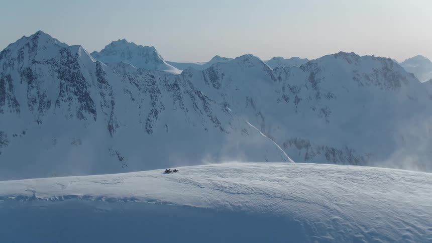 People sit on stationary snowmobiles in a snowy field near snow covered mountains on a windy winter day