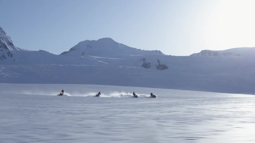 Group of four people riding snowmobiles in an environment surrounded by mountains and layers of snow