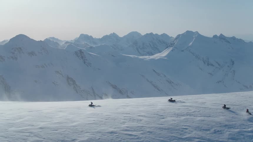 Four people are snowmobiling on a high plateau in front of a mountain range