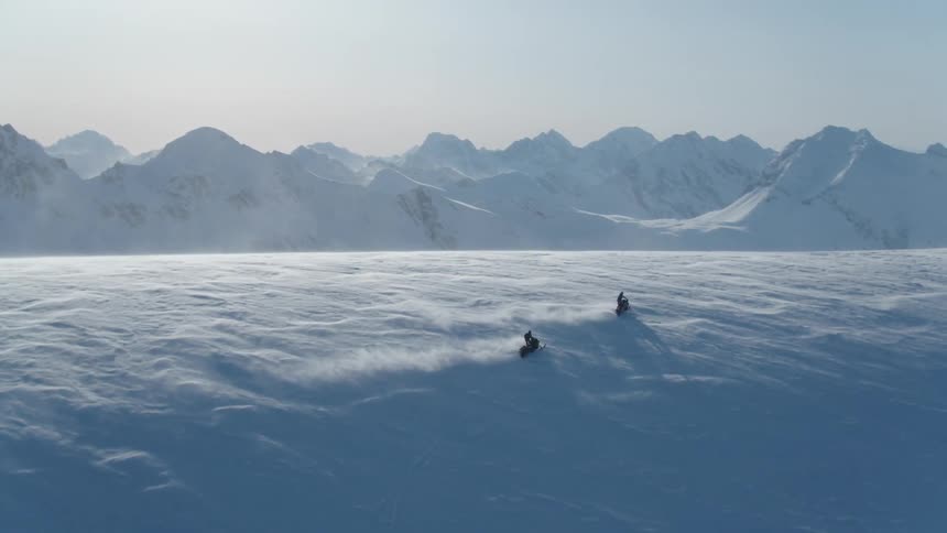People ride snowmobiles up a slope across a narrow valley from jagged snowy mountains on a sunny winter day