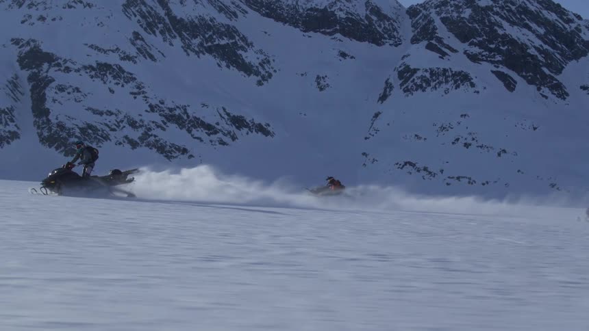 People riding snowmobiles on snowy terrain with mountains in the background