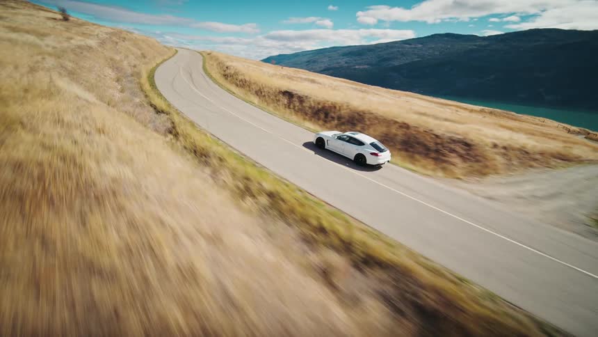 White car traveling down a road surrounded by a calm lake and mountains