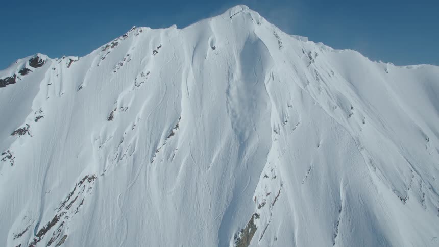 Snow falls down the side of a mountain under a clear sky