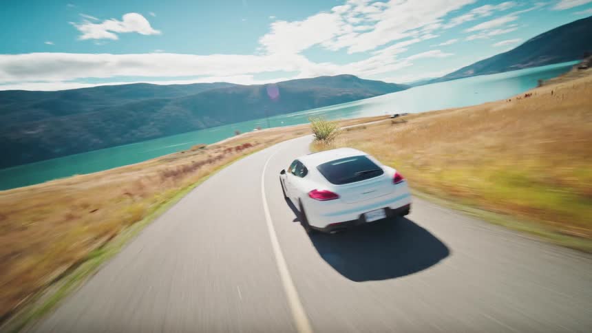 Aerial view of a white sedan sports car traveling along a road along a lake and mountains in a natural landscape