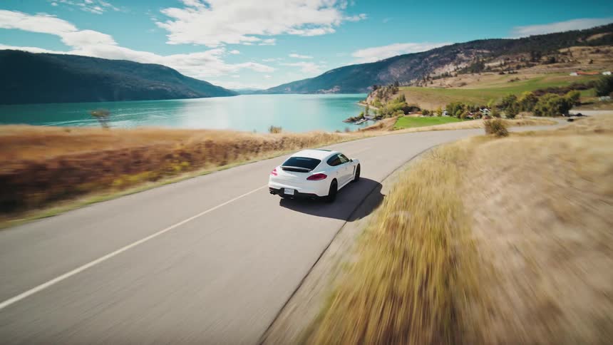 White car driving on a road next to a body of water with mountains in the background and clear sky above