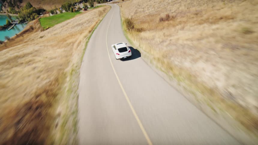 Aerial view of a white sports car traveling along a lakeside road and mountains