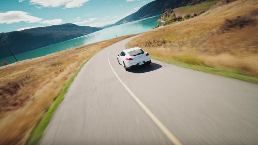 A white sports car drives along a winding road overlooking a lake