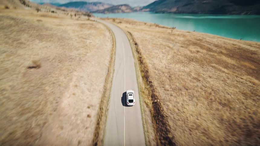 Car driving along a winding road next to a large body of water with hills in the background
