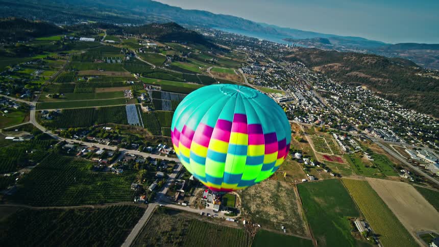 A hot air balloon drifts over a patchwork of agricultural fields and a town nestled among rolling hills