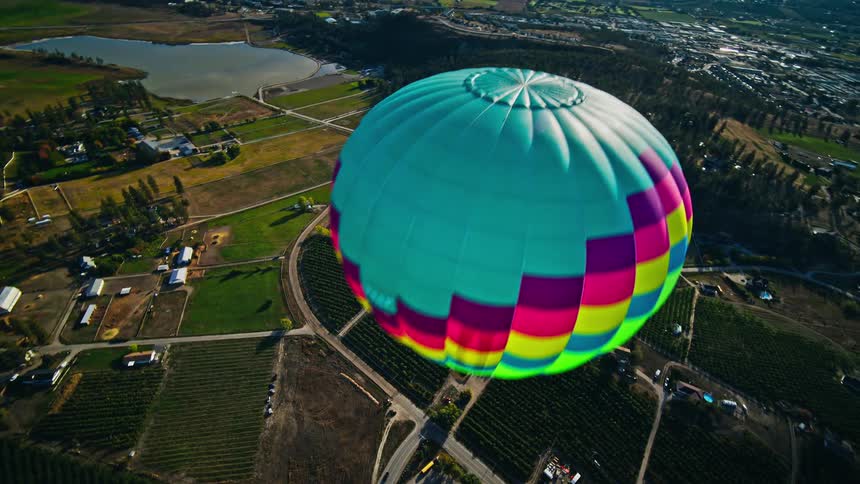 A colorful hot air balloon floats above a rural landscape with vineyards and orchards