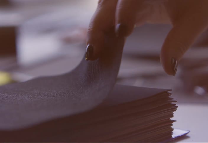 A hand carefully lifts the top sheet from a stack of black fabric revealing the layers beneath
