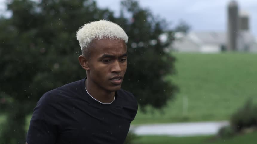 A man with white hair wearing a black shirt jogs through a green field during a rainstorm