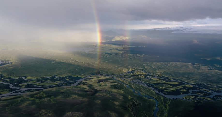 Aerial view of a vast landscape with a winding river under a sky with sunbeams and a visible rainbow