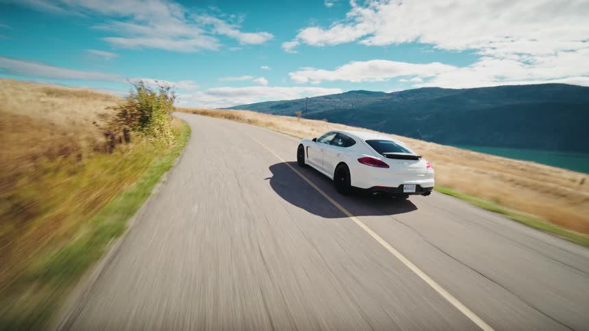 A sleek white porsche panamera cruises along a winding road with breathtaking mountain views and a shimmering lake stretching out in the distance