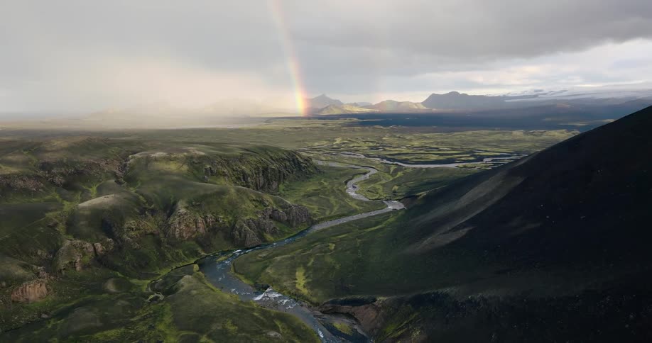 Aerial view of a vast landscape with a meandering river flanked by mountains and hills under a dramatic sky with a rainbow